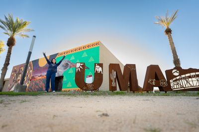 An NAU student makes a Y with her arms in the air next to a sign to spell out Yuma.