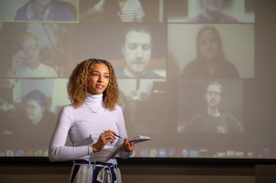 Business student gives a presentation with on-video participants on a projector screen in the background.