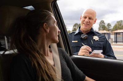 A smiling police officer standing outside a vehicle, speaking with a woman in the driver's seat while taking notes.